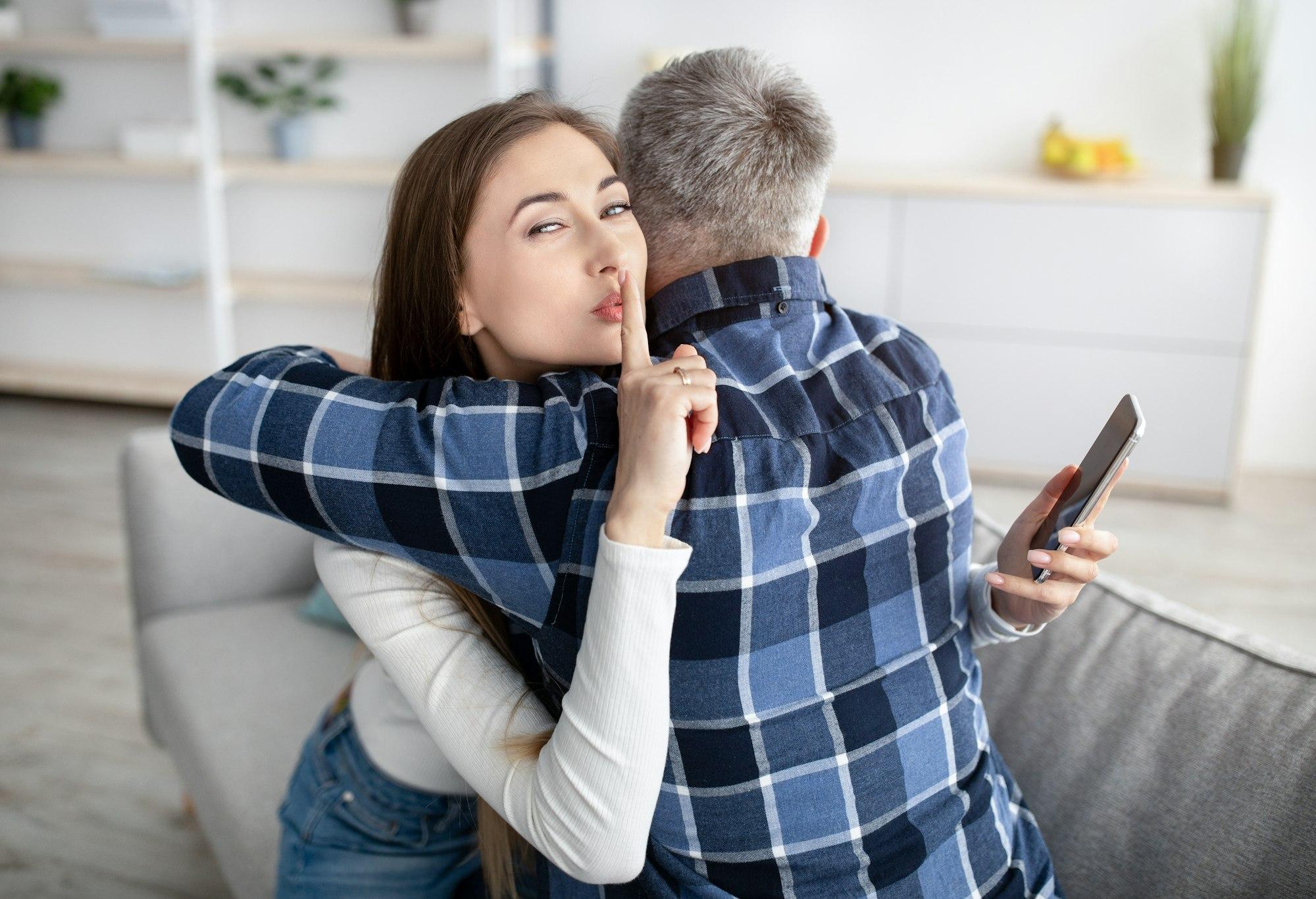 Beautiful mature woman hugging her husband, showing HUSH gesture, having secret affair, texting her