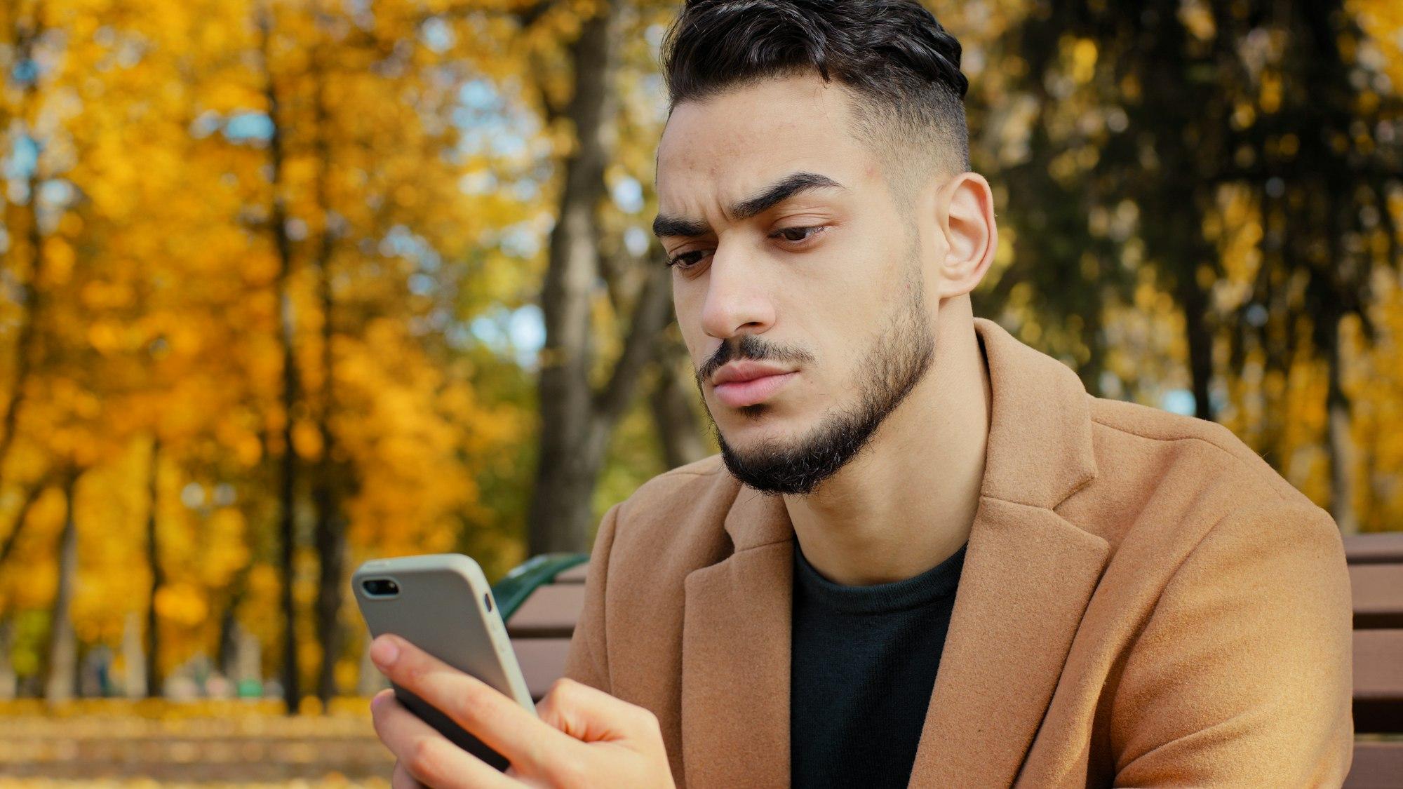 Portrait young serious guy use app hispanic man looking at phone screen browses news on telephone