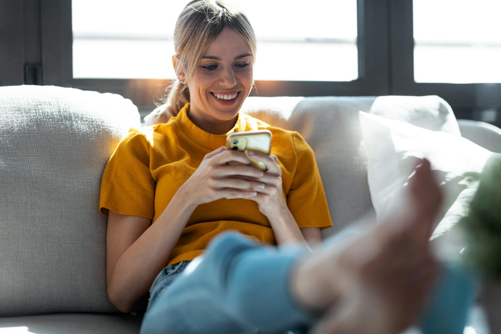 Pretty young smiling woman using mobile phone sitting on a couch at home