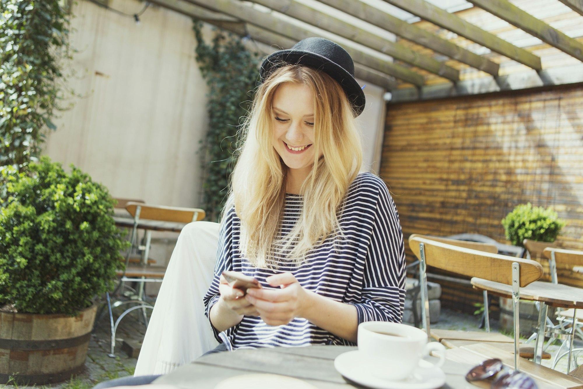 Smiling young woman wearing hat using phone while having coffee at cafe